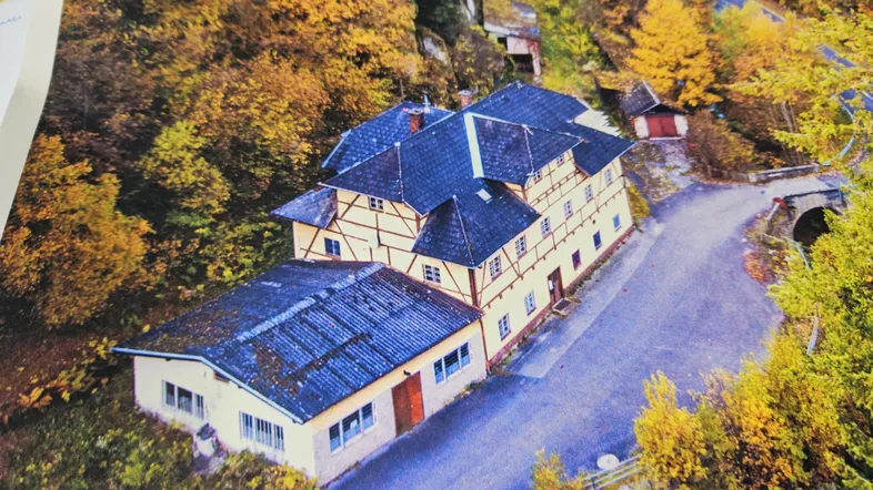 Luftaufnahme eines Gebäudekomplexes mit mehreren Dächern, umgeben von herbstlichen Bäumen und einer Straße.
