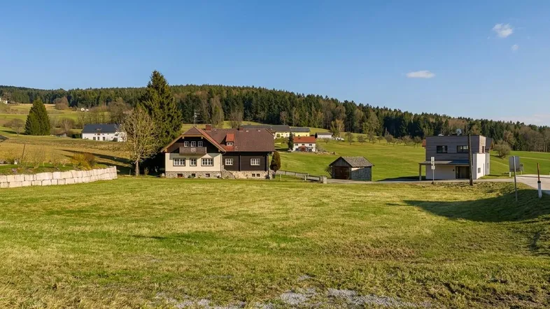 Weitläufige grüne Landschaft mit mehreren Häusern und bewaldeten Hügeln unter blauem Himmel.