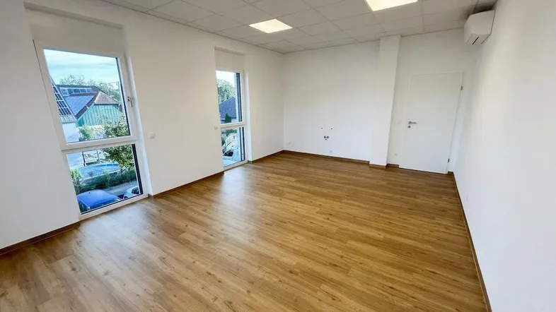 Heller Büroraum mit Laminatboden, großen Fenstern und integrierter Klimaanlage an der Wand.