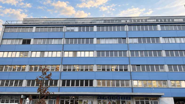 Blaues Bürogebäude mit vielen Fenstern und Geschäften im Erdgeschoss unter einem bewölkten Himmel.