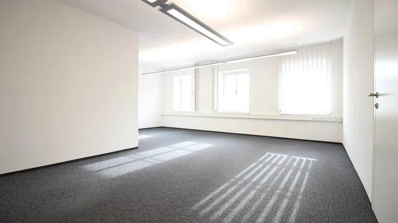 Leerer Büroraum mit Teppichboden, großen Fenstern und viel natürlichem Licht, ideal für flexible Arbeitsplatzgestaltung.