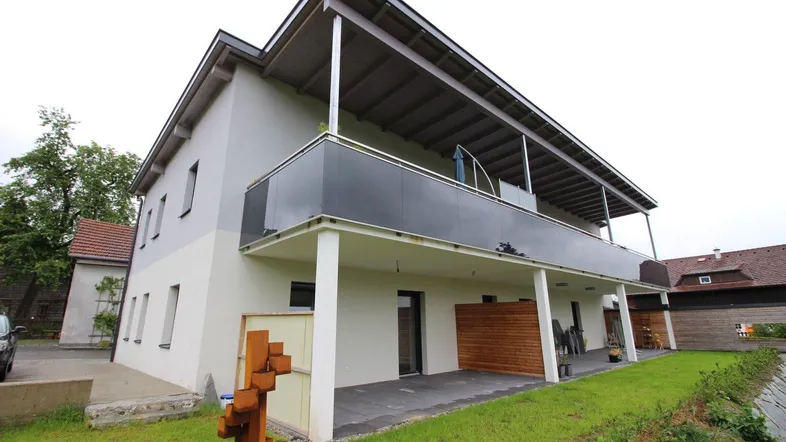 Modernes Gebäude mit Balkonen und einer gepflasterten Terrasse im Erdgeschoss, umgeben von Grünflächen.