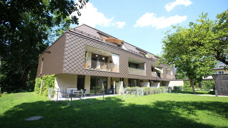 Modernes Mehrfamilienhaus mit braunem Schindeldach, Balkonen und einer Grünfläche.