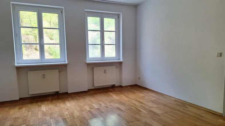 Heller, leerer Raum mit Parkettboden und zwei Fenstern, die einen Blick ins Grüne bieten.