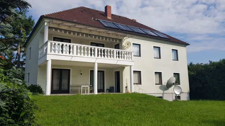 Großzügiges Wohnhaus mit Balkon, Terrasse und Solaranlage auf dem Dach, umgeben von Grünflächen.