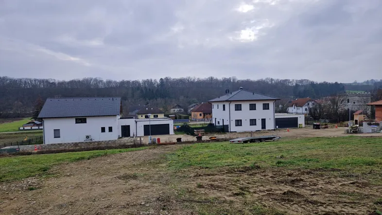 Blick auf zwei moderne Neubauten in ländlicher Umgebung mit unbebautem Grundstück im Vordergrund.