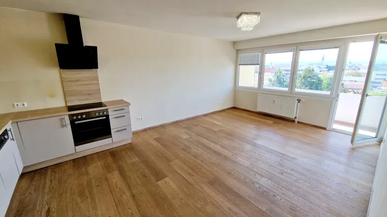 Heller offener Wohnbereich mit integrierter Küche, Parkettboden und Zugang zum Balkon.
