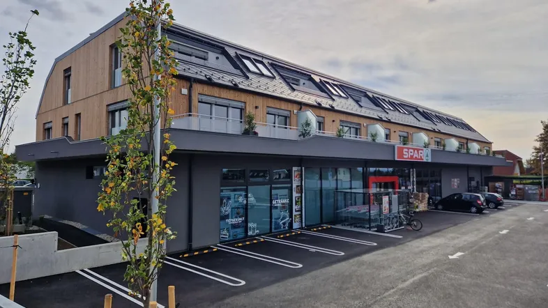 Modernes Gebäude mit Holzfassade und integriertem Supermarkt im Erdgeschoss, umgeben von Parkplätzen.