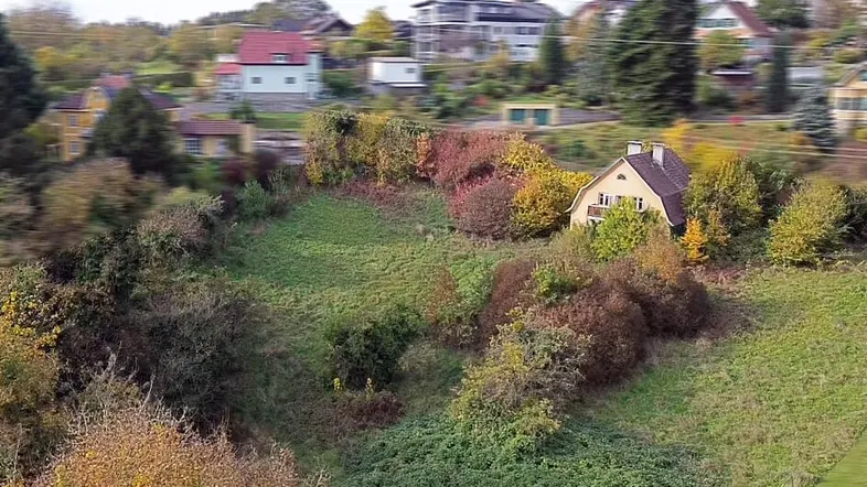 Luftaufnahme eines Hauses auf einem bewachsenen Hanggrundstück mit Herbstlaub und weiteren Häusern im Hintergrund.