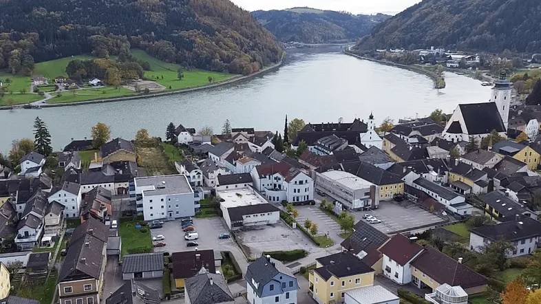 Luftaufnahme einer malerischen Stadt am Flussufer, umgeben von bewaldeten Hügeln und Wohngebäuden.