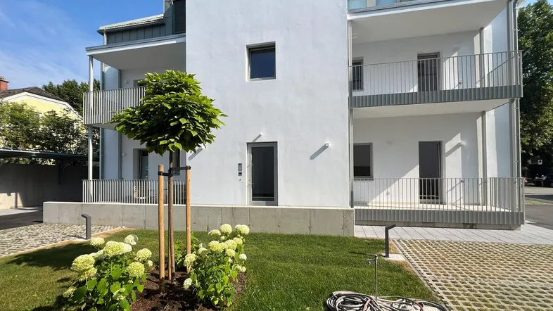 Modernes Wohngebäude mit weißer Fassade, Balkonen und gepflegtem Vorgarten an einem sonnigen Tag.