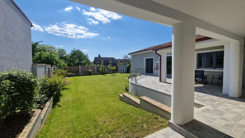 Überdachte Terrasse mit Fliesenboden und Blick auf den gepflegten Garten unter blauem Himmel.