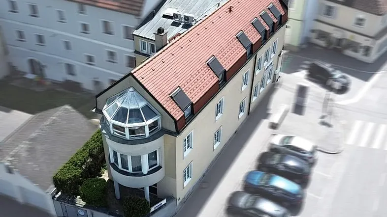 Luftaufnahme eines modernen Gebäudes mit rotem Ziegeldach und markantem Erker, umgeben von Parkplätzen und Hecken.