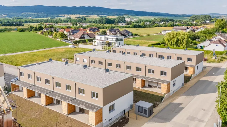 Luftaufnahme von modernen Reihenhäusern in ländlicher Umgebung mit viel Grün und Bergen im Hintergrund.