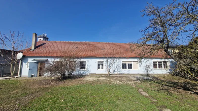 Langes, hellblaues Gebäude mit rotem Ziegeldach und mehreren Fenstern, umgeben von einem ungepflegten Garten.