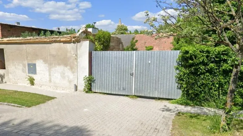 Ansicht der Gebäudefassade mit Putz, einem Metalltor und üppiger Hecke im Vorgarten.