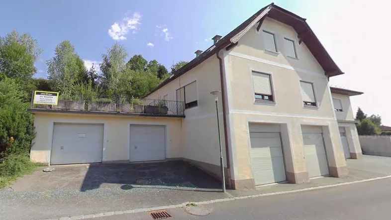 Mehrfamilienhaus mit mehreren Garagen und Balkon an sonnigem Tag, umgeben von Bäumen.