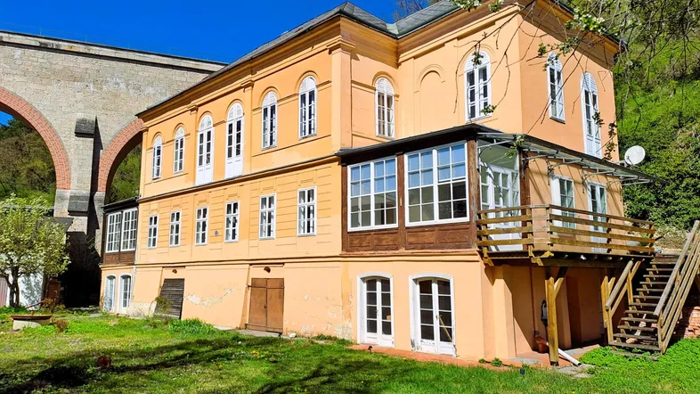 Historisches Gebäude mit gelber Fassade und Viadukt im Hintergrund, umgeben von viel Grün.
