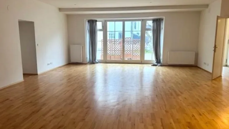 Helles, leeres Wohnzimmer mit Holzboden und großen Fenstern, die zu einem Außenbereich führen.