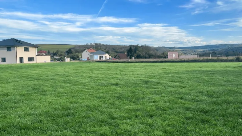 Weitläufiges grünes Feld mit Häusern im Hintergrund und hügeliger Landschaft unter blauem Himmel.