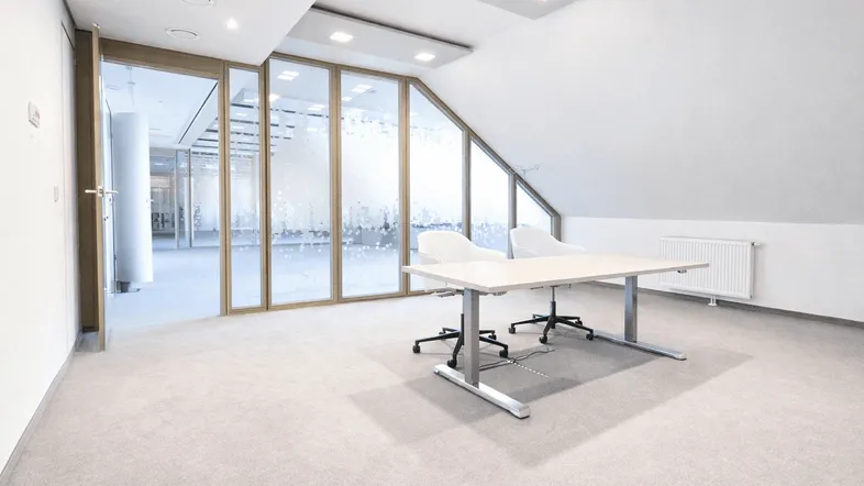 Geräumiges Büro mit Teppichboden, schrägen Wänden und großen Glasfronten zu weiteren Büroräumen.