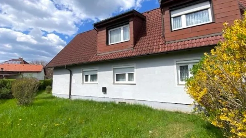 Hausfassade mit rotem Ziegeldach, Gaubenfenstern und grünem Vorgarten unter blauem Himmel.