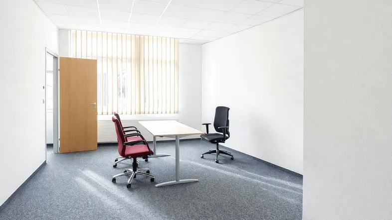Helles Büro mit Teppichboden, vertikalen Jalousien an großen Fenstern und einfacher Möblierung.