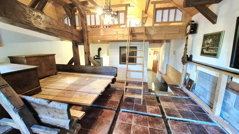 Geräumiger Wohnbereich mit sichtbaren Holzbalken, rustikalen Fliesen und einer Holzleiter zum oberen Bereich.