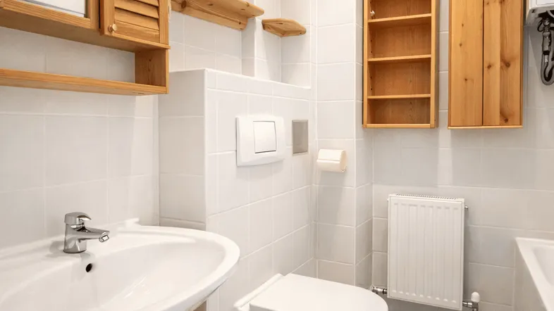 Helles Badezimmer mit weißen Fliesen, Holzschränken, Waschbecken, Toilette und Heizkörper.