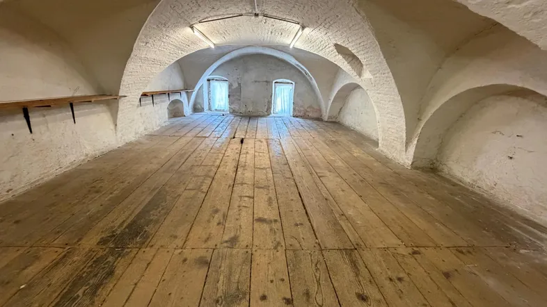 Geräumiger Lagerraum mit gewölbter Decke und rustikalem Holzboden, beleuchtet durch kleine Fenster.