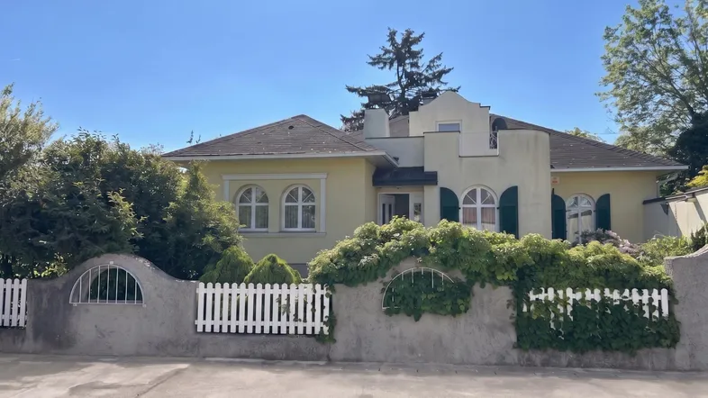 Gelbe Villa mit weißen Fensterrahmen, grünen Fensterläden und einem gepflegten Vorgarten.