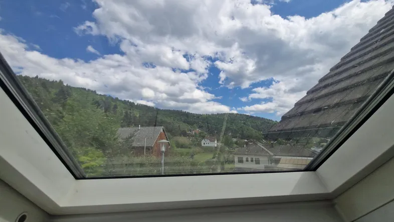 Blick aus einem Dachfenster auf eine grüne Hügellandschaft mit Häusern und bewölktem Himmel.