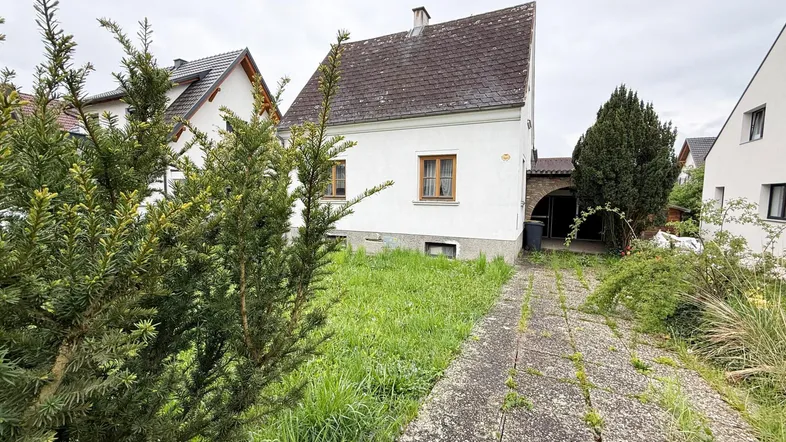 Weißes Haus mit Satteldach und verwachsenem Vorplatz, umgeben von weiteren Gebäuden.