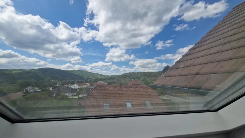 Panoramablick aus einem Dachfenster über Dächer, grüne Hügel und eine weite Landschaft.