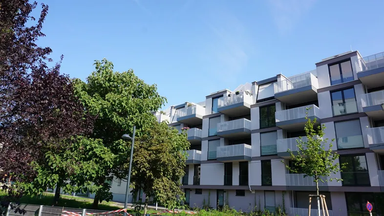 Modernes Wohngebäude mit mehreren Balkonen und viel Grün in der Umgebung unter blauem Himmel.
