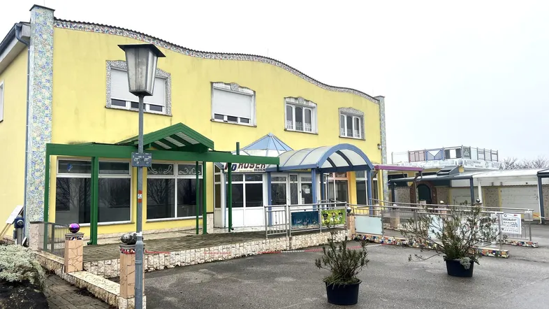 Gelbes Gebäude mit geschwungener Fassade, mehreren Eingängen und Vordächern. Eine Rampe führt zum Eingangsbereich, davor befindet sich ein Parkplatz.