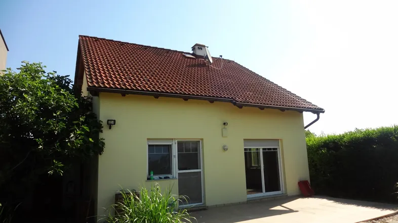 Helles Einfamilienhaus mit Ziegeldach, gelber Fassade und einer kleinen gepflasterten Terrasse im Garten.