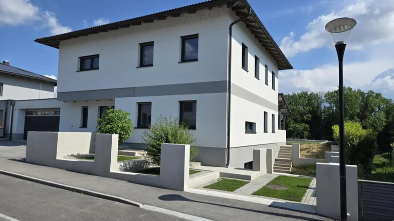 Modernes Einfamilienhaus mit weißer Fassade, grauen Akzenten und gepflegter Außenanlage unter blauem Himmel.