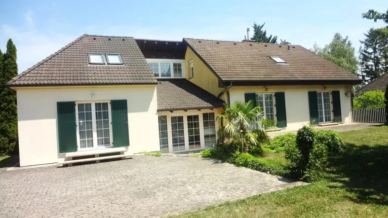 Großzügiges Einfamilienhaus mit heller Fassade, grünen Fensterläden und gepflasterter Auffahrt.
