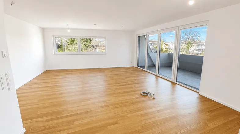 Helles, leeres Zimmer mit Holzboden, großen Fenstern und Zugang zum Balkon.