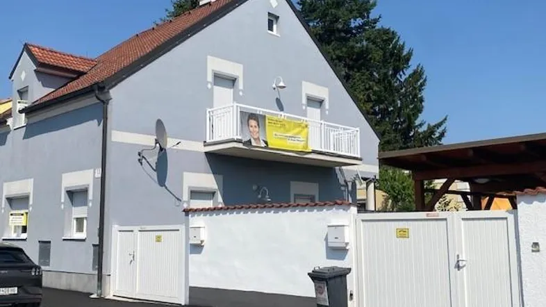 Einfamilienhaus mit hellblauer Fassade, Satteldach und kleinem Balkon, umgeben von einem Zaun und einem Carport.