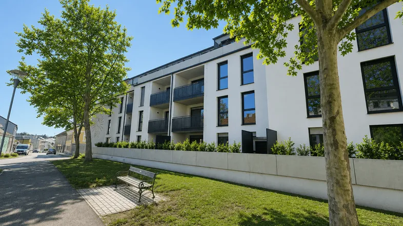 Modernes Mehrfamilienhaus mit weißer Fassade, Balkonen und gepflegter Grünanlage an einem sonnigen Tag.