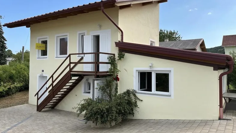 Gelbes Haus mit Außentreppe und gepflastertem Vorplatz unter blauem Himmel.