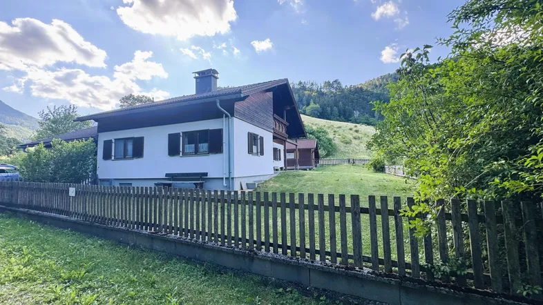 Traditionelles Haus mit Holzzaun und grüner Umgebung unter blauem Himmel mit Wolken.