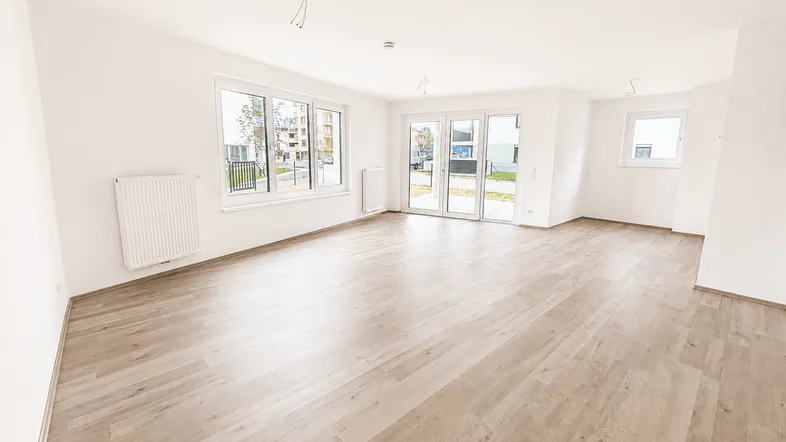 Helles, leeres Wohnzimmer mit Laminatboden und großen Fenstern, die viel Tageslicht hereinlassen.