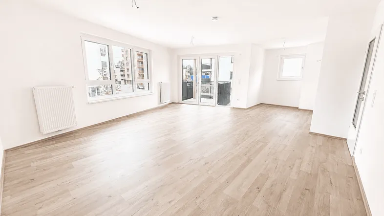 Helles, leeres Wohnzimmer mit Laminatboden, großen Fenstern und Zugang zu einem Balkon.