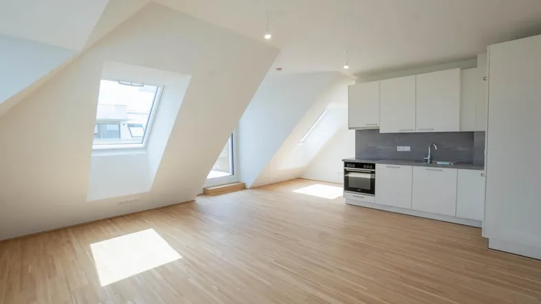 Heller Dachgeschossraum mit kleiner Einbauküche, Holzparkett und schrägen Wänden mit Dachfenstern.