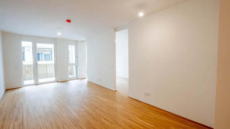 Helles, leeres Zimmer mit Holzboden und Zugang zu einem Balkon, bereit zur Einrichtung.
