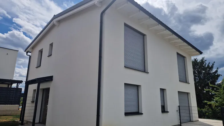 Modernes Einfamilienhaus mit weißer Fassade, dunklem Dach und Rollläden an den Fenstern.