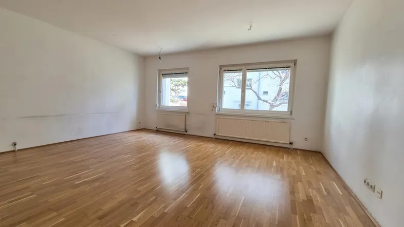 Helles, leeres Zimmer mit Parkettboden und zwei Fenstern, bereit zur individuellen Gestaltung.